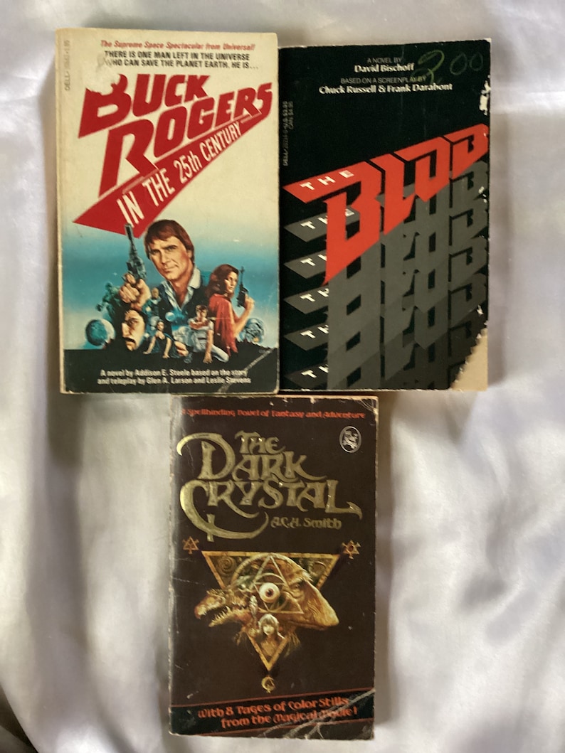 May include: Three paperback books, one with a red and black cover featuring the text "The Blood", one with a red and white cover featuring the text "Buck Rogers in the 25th Century", and one with a brown cover featuring the text "The Dark Crystal" and a graphic of a creature with a large eye.