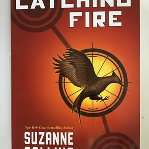 May include: The cover of the book "Catching Fire" by Suzanne Collins, the second book in the Hunger Games trilogy. The cover features a red background with a stylized image of a mockingjay in flight, surrounded by a yellow circle with a target design. The text "Catching Fire" is in white letters, and the author's name, "Suzanne Collins", is in white letters below the image.