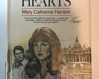 May include: A book cover with a romantic novel title, "Captured Hearts" by Mary Catherine Hanson. The cover features a woman with long brown hair and a man with short brown hair. The background is a drawing of a building with a dome.