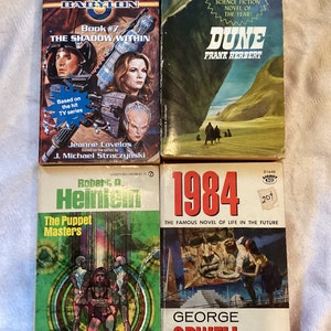 May include: Four paperback science fiction novels. The first is "Babylon 5: The Shadow Within" by Jeanne Cavelos and J. Michael Straczynski. The second is "Dune" by Frank Herbert. The third is "The Puppet Masters" by Robert A. Heinlein. The fourth is "1984" by George Orwell.
