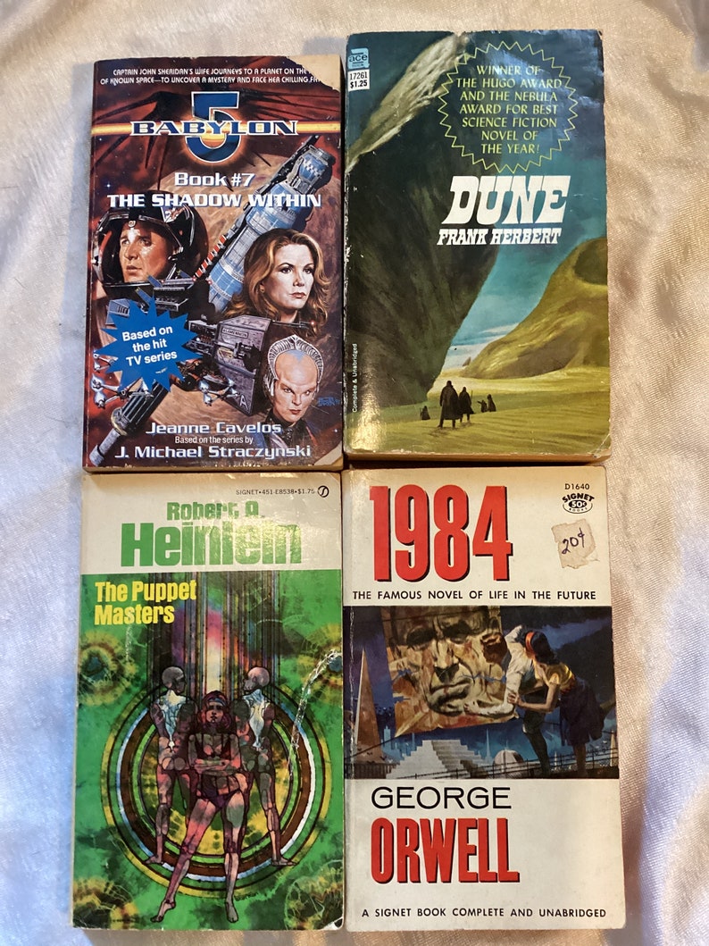 May include: Four paperback science fiction novels. The first is "Babylon 5: The Shadow Within" by Jeanne Cavelos and J. Michael Straczynski. The second is "Dune" by Frank Herbert. The third is "The Puppet Masters" by Robert A. Heinlein. The fourth is "1984" by George Orwell.