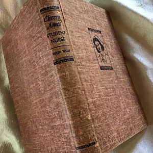 May include: A brown book with the title "Cherry Ames Student Nurse" by Helen Wells. The book cover features a black and white illustration of a woman in a nurse's uniform.