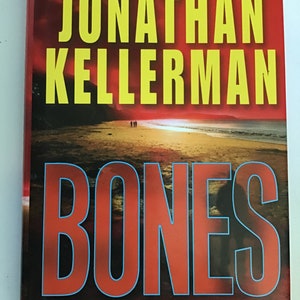 May include: A book cover with a red background and the title "Bones" in large, bold, red letters with a blue outline. The author's name, "Jonathan Kellerman", is printed in yellow above the title. The book is described as "An Alex Delaware Novel".