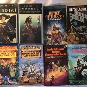 May include: A collection of ten paperback fantasy and science fiction novels. The books are arranged in two rows of five. The top row features books with titles like "Abhorsen", "Retief of the CDT", and "Rogue Bolo". The bottom row features books with titles like "Slaves of the Volcano God", "Revenge of the Fluffy Bunnies", "Lando Calrissian and the Flamewind of Oscon", and "Brightsuit Macbear".