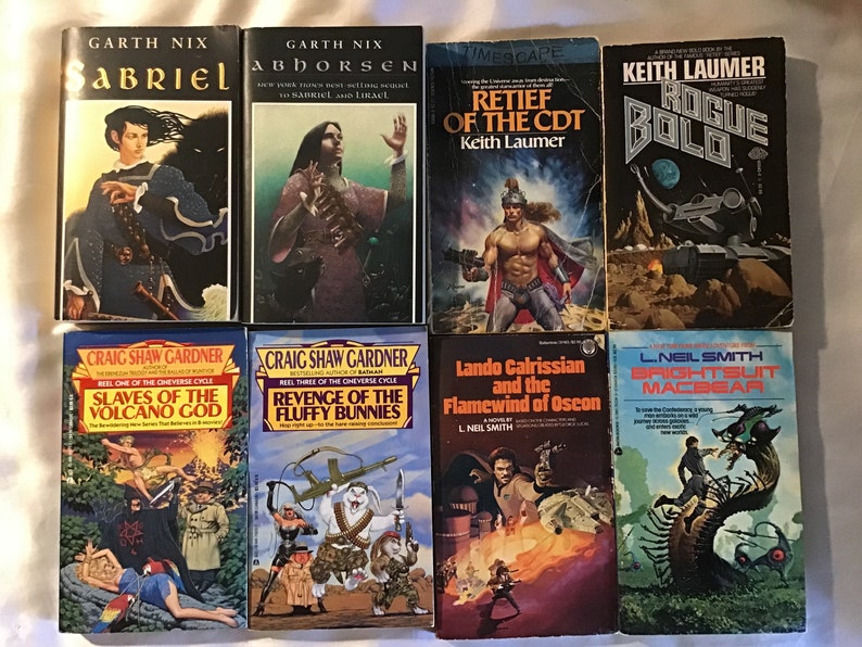 May include: A collection of ten paperback fantasy and science fiction novels. The books are arranged in two rows of five. The top row features books with titles like "Abhorsen", "Retief of the CDT", and "Rogue Bolo". The bottom row features books with titles like "Slaves of the Volcano God", "Revenge of the Fluffy Bunnies", "Lando Calrissian and the Flamewind of Oscon", and "Brightsuit Macbear".
