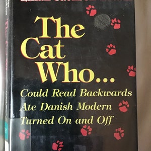 May include: A black book cover with the title "The Cat Who... Could Read Backwards Ate Danish Modern Turned On and Off" in yellow and red lettering. The book cover also features the text "Lilian Jackson Braun" and "Three Novels Complete and Unabridged".
