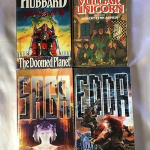 May include: A collection of four paperback books. The top book is titled "The Doomed Planet" by L. Ron Hubbard. The second book is titled "Tales from the Vulgar Unicorn" edited by Robert Lynn Asprin. The third book is titled "Saga" by Conor Kostick. The fourth book is titled "Edda" by Conor Kostick.