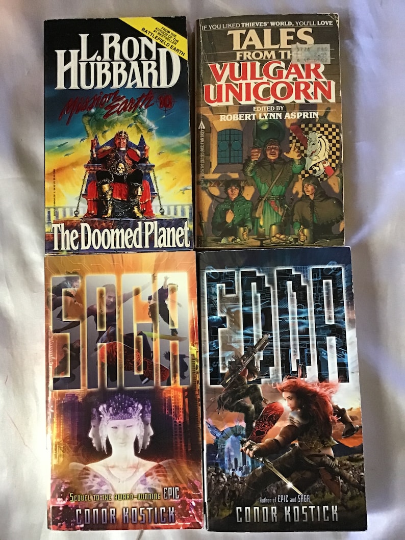 May include: A collection of four paperback books. The top book is titled "The Doomed Planet" by L. Ron Hubbard. The second book is titled "Tales from the Vulgar Unicorn" edited by Robert Lynn Asprin. The third book is titled "Saga" by Conor Kostick. The fourth book is titled "Edda" by Conor Kostick.
