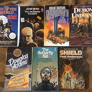 May include: A collection of ten paperback science fiction and fantasy novels. The books are arranged in two rows, with five books in each row. The books have colorful covers with illustrations and text. The titles of the books include "The Eye of the World", "The Last Starship from Earth", "Voyagers II", "Demon Undoing", "Dirk Gently's Holistic Detective Agency", "The Butterfly Kid", and "Shield".