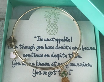 Puede incluir: Un brazalete dorado con un dije de piña y un pequeño dije de corazón blanco. El brazalete está en una caja de regalo turquesa con una tarjeta que dice "¡Sé imparable! Aunque tengas dudas y miedos, continúa a pesar de todo. Eres un guerrero valiente y fuerte. ¡Tú puedes!"