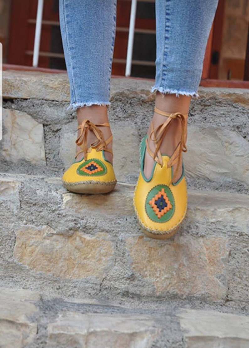 Puede incluir: Sandalias de cuero amarillas con costuras decorativas y un dise&ntilde;o geom&eacute;trico en verde, naranja y azul marino. Las sandalias tienen un lazo en el tobillo y se usan con vaqueros azules.