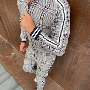 May include: A matching plaid tracksuit in gray, black, and burgundy. The jacket has a black collar and cuffs with white stripes, and white stripes run down the sleeves. The pants have white stripes down the sides. The person is wearing white sneakers.