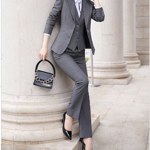 May include: A gray women's suit with a blazer, vest, and pants. The suit is tailored and has a professional look. The woman is wearing black heels and a white shirt with a black tie.