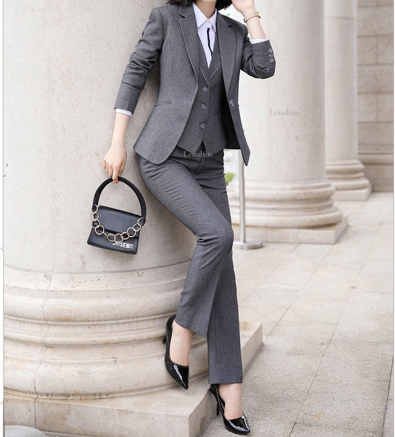 May include: A gray women's suit with a blazer, vest, and pants. The suit is tailored and has a professional look. The woman is wearing black heels and a white shirt with a black tie.