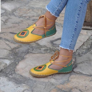 Puede incluir: Sandalias de cuero amarillas y verdes con un dise&ntilde;o decorativo en la puntera. Las sandalias tienen un dise&ntilde;o con cordones con cordones de color beige. Las sandalias se usan con vaqueros azules.