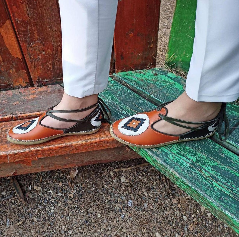 Puede incluir: Sandalias de cuero marr&oacute;n y negro con un dise&ntilde;o &uacute;nico. Las sandalias presentan un patr&oacute;n decorativo en la puntera y una correa que rodea el tobillo. Las sandalias se muestran sobre un banco de madera. Un calzado &uacute;nico.