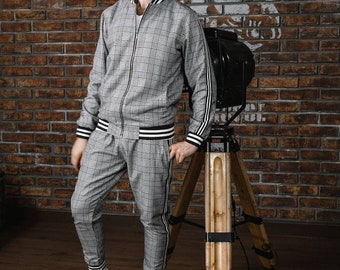 May include: A gray plaid tracksuit with white and black striped accents. The jacket has a zipper and the pants are fitted. The outfit is paired with white sneakers and black sunglasses. The background is a brick wall.