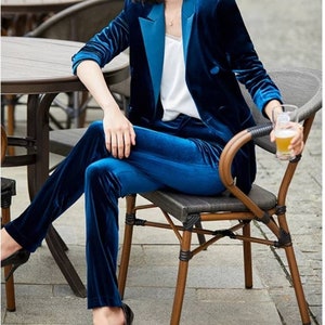 May include: A two-piece velvet suit in a deep blue hue. The suit includes a blazer with a contrasting lapel and matching trousers. The outfit is paired with a white top. The model is seated on a chair, wearing black heels.