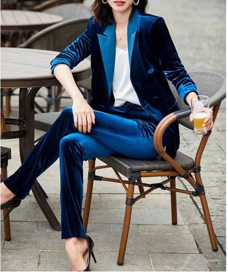 May include: A two-piece velvet suit in a deep blue hue. The suit includes a blazer with a contrasting lapel and matching trousers. The outfit is paired with a white top. The model is seated on a chair, wearing black heels.