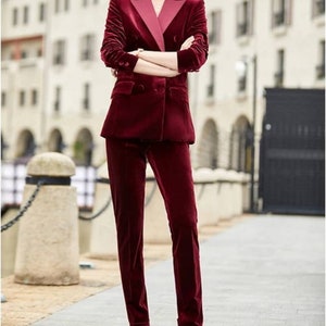 May include: A burgundy velvet suit, featuring a double-breasted blazer with a contrasting pink lapel and matching straight-leg trousers. The suit is worn with black heels. The outfit is a sophisticated ensemble.