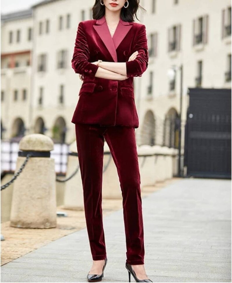 May include: A burgundy velvet suit, featuring a double-breasted blazer with a contrasting pink lapel and matching straight-leg trousers. The suit is worn with black heels. The outfit is a sophisticated ensemble.
