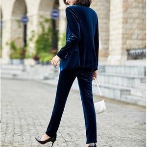 May include: A teal velvet pantsuit. The blazer has a notched lapel and button closure. The pants are straight-legged. The suit is paired with black heels and a white purse with a gold chain. The outfit is worn on a cobblestone street.