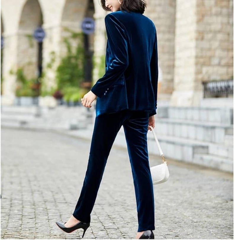 May include: A teal velvet pantsuit. The blazer has a notched lapel and button closure. The pants are straight-legged. The suit is paired with black heels and a white purse with a gold chain. The outfit is worn on a cobblestone street.