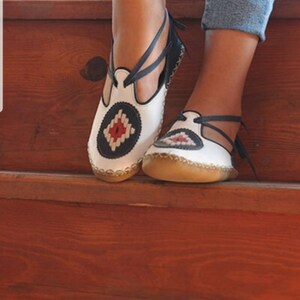Puede incluir: Sandalias de cuero blanco con un dise&ntilde;o decorativo en azul marino y rojo. Las sandalias tienen una correa que se envuelve alrededor del tobillo. Las suelas son de color marr&oacute;n claro. Sobre una superficie de madera.