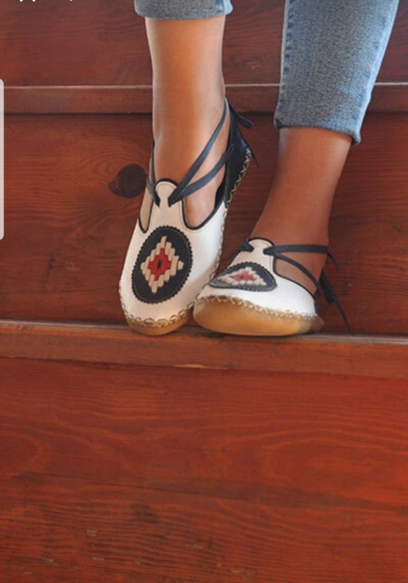Puede incluir: Sandalias de cuero blanco con un dise&ntilde;o decorativo en azul marino y rojo. Las sandalias tienen una correa que se envuelve alrededor del tobillo. Las suelas son de color marr&oacute;n claro. Sobre una superficie de madera.