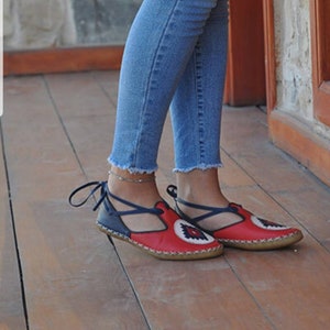 Puede incluir: Bailarinas de cuero rojas y azul marino con un dise&ntilde;o floral decorativo. Las bailarinas tienen una correa que se envuelve alrededor del tobillo y se usan con vaqueros azules.