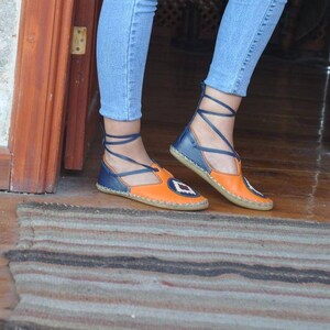 Puede incluir: Sandalias de cuero naranja y azul con un dise&ntilde;o geom&eacute;trico en la puntera. Las sandalias tienen una correa de tobillo con cordones y una suela plana. Los zapatos se usan con vaqueros azules.