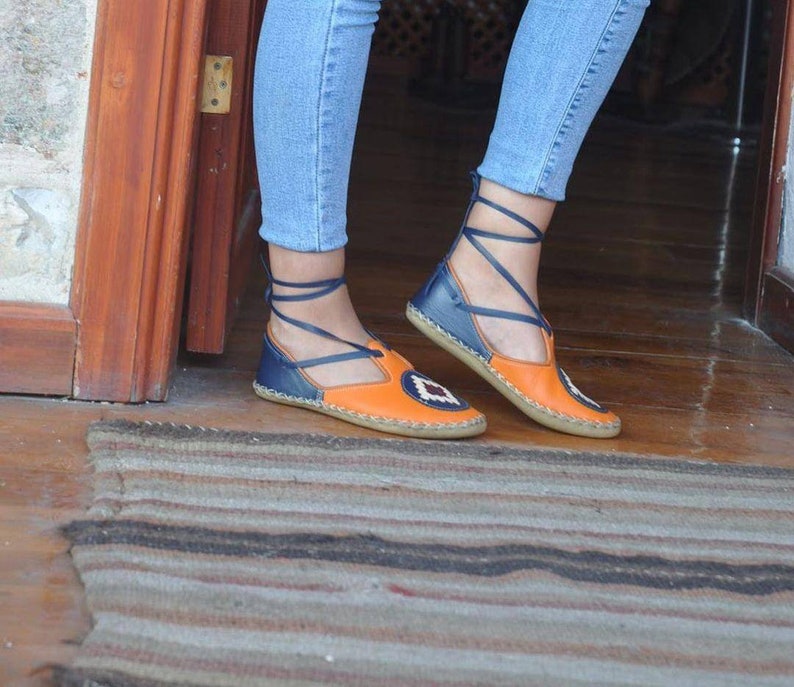 Puede incluir: Sandalias de cuero naranja y azul con un dise&ntilde;o geom&eacute;trico en la puntera. Las sandalias tienen una correa de tobillo con cordones y una suela plana. Los zapatos se usan con vaqueros azules.