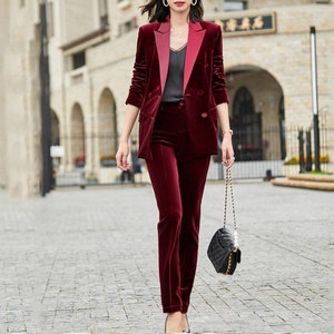 May include: A burgundy velvet pantsuit with a contrasting lapel on the blazer. The double-breasted blazer and matching trousers create a coordinated ensemble. A black quilted handbag with a chain strap completes the outfit.