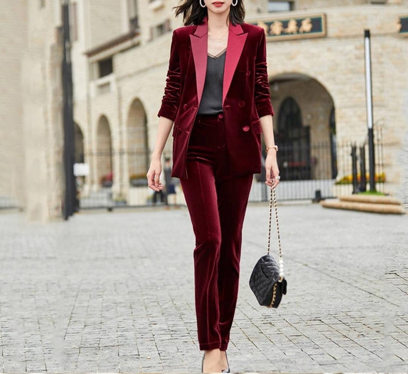 May include: A burgundy velvet pantsuit with a contrasting lapel on the blazer. The double-breasted blazer and matching trousers create a coordinated ensemble. A black quilted handbag with a chain strap completes the outfit.