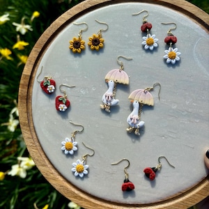 May include: A collection of handmade earrings featuring various designs, including sunflowers, strawberries, daisies, ladybugs, and geese with umbrellas. The earrings are displayed on a round, light gray surface with a wooden border, set against a backdrop of green foliage.