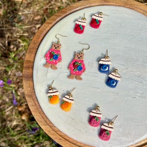 May include: A collection of handmade polymer clay earrings. The earrings feature various designs, including bear figures in pink sweaters, fruit-filled jars, and berry-topped cups. The earrings are displayed on a light blue surface.