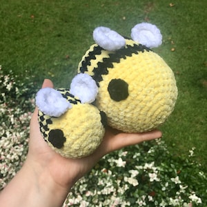 May include: Two plush, handmade bee toys. The bees are yellow with black stripes and black eyes. They have white fuzzy wings. The larger bee is approximately 6 inches in diameter, and the smaller bee is about 3 inches in diameter. The bees are held in a hand.