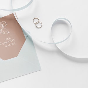 May include: A light blue envelope with a peach-coloured card inside. The card features a white line drawing of a flower and the text "Save the Date 10.12.2022". Two gold rings are on a white surface next to the envelope.
