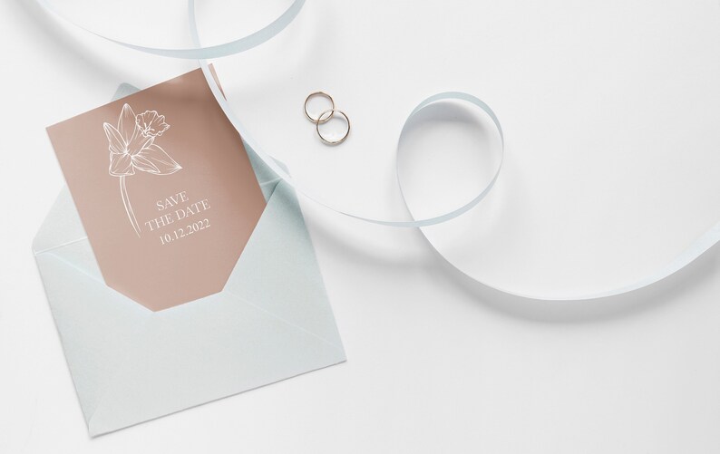 May include: A light blue envelope with a peach-coloured card inside. The card features a white line drawing of a flower and the text "Save the Date 10.12.2022". Two gold rings are on a white surface next to the envelope.