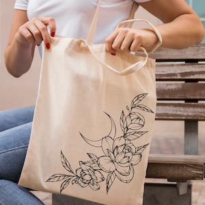 May include: A beige canvas tote bag with a black floral design featuring a crescent moon and leaves. The design is in the lower right corner of the bag.