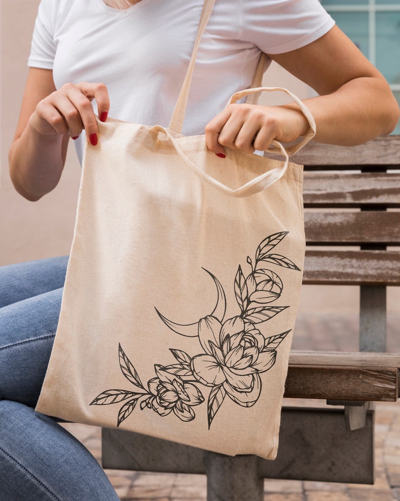 May include: A beige canvas tote bag with a black floral design featuring a crescent moon and leaves. The design is in the lower right corner of the bag.