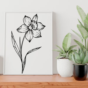 May include: A black and white line drawing of a daffodil flower with a single stem and leaves. The drawing is framed and displayed on a white wall above a wooden table.