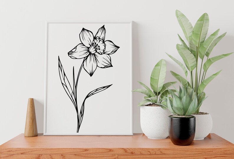 May include: A black and white line drawing of a daffodil flower with a single stem and leaves. The drawing is framed and displayed on a white wall above a wooden table.