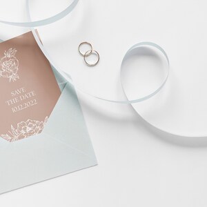 May include: A light blue envelope with a peach-colored card inside. The card has a floral design and the text "Save the Date 10.12.2022". Two gold rings are on a white surface next to the envelope.