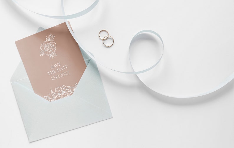 May include: A light blue envelope with a peach-colored card inside. The card has a floral design and the text "Save the Date 10.12.2022". Two gold rings are on a white surface next to the envelope.