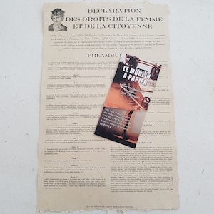 Könnte beinhalten: Ein altes Dokument mit dem Titel "Deklaration der Rechte der Frau und der Bürgerin" mit einem französischen Text über Frauenrechte. Das Dokument ist auf gealtertem Papier gedruckt und hat ein verblasstes Aussehen. Ein kleines, gefaltetes Pamphlet für eine Papiermühle ist in der rechten unteren Ecke sichtbar.