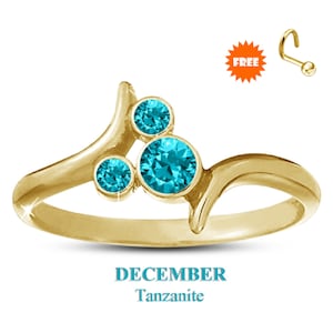 May include: A gold-colored ring with three round, bright blue tanzanite stones. The ring has a unique, flowing design. The word "DECEMBER" and "Tanzanite" are displayed in light blue text below the ring. A free nose ring is included.