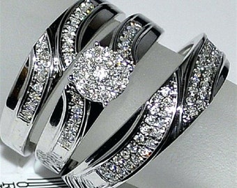 Könnte beinhalten: Ein Set aus drei silbernen Ringen. Der Verlobungsring hat einen Cluster aus kleinen, runden Diamanten. Die Bänder sind mit kleineren Diamanten entlang eines geschwungenen Designs verziert. Die Ringe werden auf einer weißen Oberfläche präsentiert.