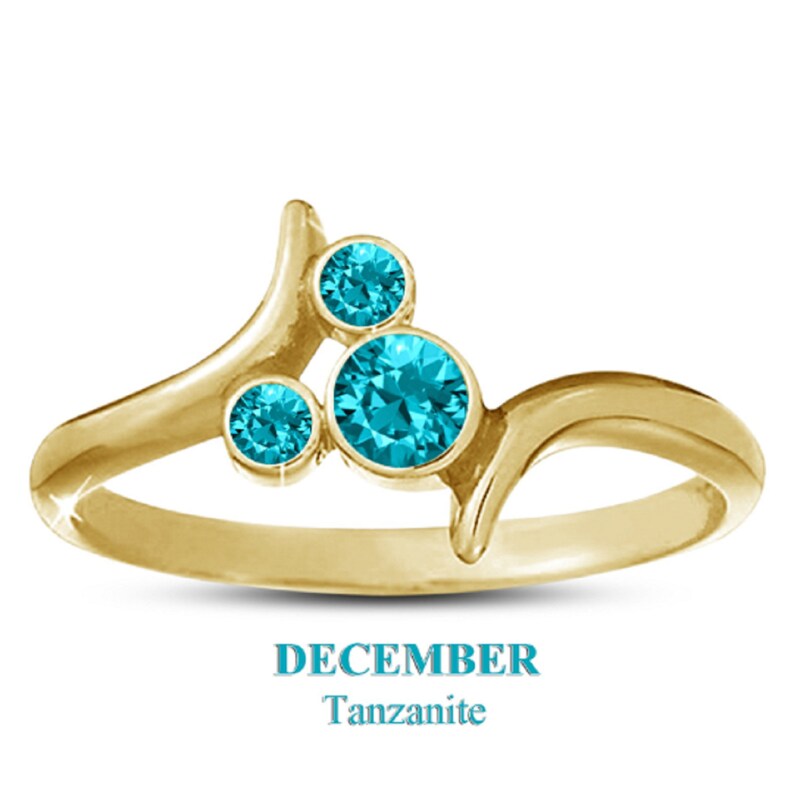 May include: A gold-colored ring with three round, bright blue gemstones. The ring has a unique, flowing design with the text "DECEMBER Tanzanite" below the ring.