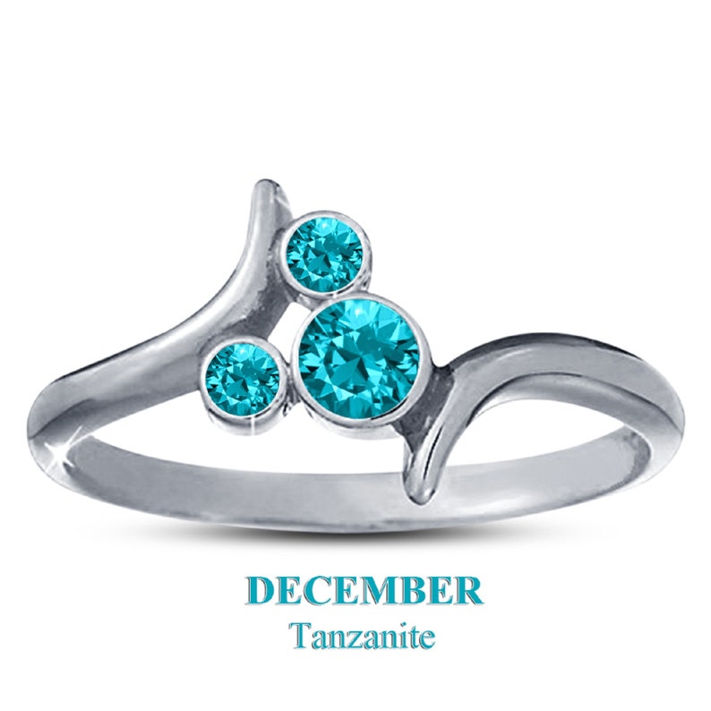 May include: A silver ring with three round, teal-colored tanzanite stones. The ring has a unique, flowing design with the text "DECEMBER Tanzanite" below the ring.
